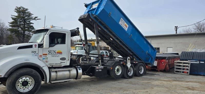A white semi-truck with the Sun Environmental Corp logo on the door uses a hydraulic system to lower a bright blue roll-off bin to the ground